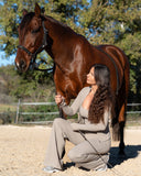 Woman in a beige outfit kneeling next to a brown horse in an outdoor setting with trees in the background.