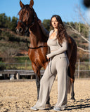 Woman in a light gray outfit standing next to a brown horse in an outdoor setting.